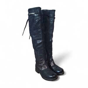 Könnte beinhalten: Ein Paar schwarze Overknee-Stiefel. Die Stiefel sind aus einem strukturierten Ledermaterial gefertigt. Jeder Stiefel hat eine silberfarbene Schnalle oben und eine Schnürung. Die Stiefel haben einen niedrigen Absatz und eine runde Spitze.