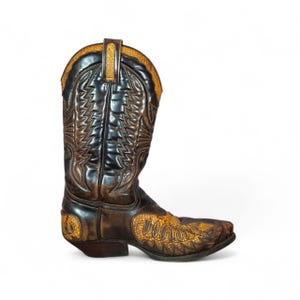 Puede incluir: Una bota de vaquero marrón y negra con intrincadas costuras y un patrón decorativo en la puntera. La bota presenta una puntera puntiaguda y un tacón apilado, mostrando un diseño occidental clásico. La bota está sobre un fondo blanco.