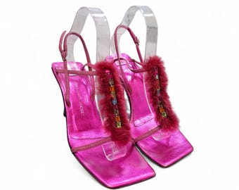 May include: A pair of metallic fuchsia high-heeled sandals featuring a square toe design, thin ankle straps with buckles, and a vertical strip of dark pink faux fur accented with a row of small, multicolored rectangular crystals on the front.