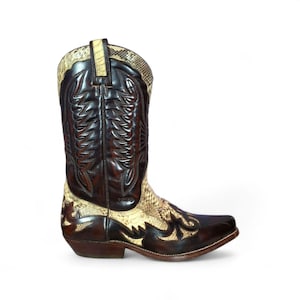 Puede incluir: Bota de vaquero de cuero marrón con puntera cuadrada. La bota presenta un estampado de piel de serpiente en la caña y la pala, con costuras decorativas. La bota tiene un tacón apilado y una trabilla.