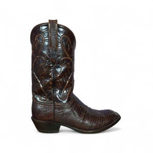 May include: A single brown cowboy boot with a pointed toe and a stacked heel. The boot has detailed embossed patterns and a textured finish, suggesting a high-quality leather construction. The boot is set against a white background.