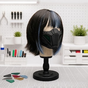 Op de afbeelding: Een zwarte pruik met blauwe highlights in een korte boblijn met pony, gedragen op een etalagepophoofd. De etalagepop draagt een matzwart, hoekig, geometrisch gezichtsmasker met groene mesh-details over de mond en neus.