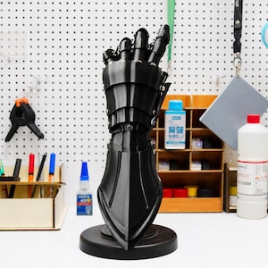 May include: A black articulated metal gauntlet standing upright on a circular base. The gauntlet features overlapping plates covering the fingers, hand, and wrist, secured with small silver-colored rivets, creating a protective, segmented structure.