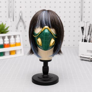 May include: A dark brown wig with blue highlights styled in a short bob, worn on a mannequin head. The mannequin wears a green and gold face mask featuring a geometric, hexagonal pattern and two prominent gold-coloured circular side vents.