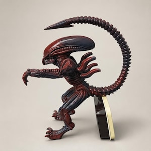 May include: A dark reddish-brown xenomorph figurine featuring an elongated head, sharp claws, and a long, curved tail that arches over its back. The creature is depicted in a crouched, aggressive stance with its arms extended forward.