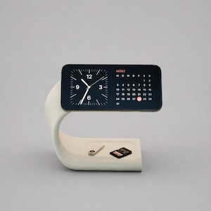 May include: A light gray curved concrete phone stand holding a smartphone displaying a clock and a calendar for MÄRZ. On the base of the stand sits a small metallic ring and a black memory card. The stand has a smooth, matte finish.
