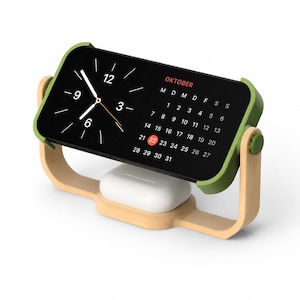 Op de afbeelding: Een smartphone in een groene houder op een licht houtkleurige standaard, met een zwarte digitale klok en een kalender voor de maand OKTOBER waarbij de 22e in het rood is gemarkeerd. Een witte oplaadcase staat op de basis van de standaard.