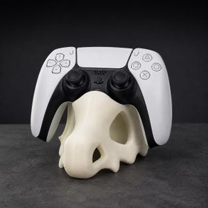 Könnte beinhalten: Ein weiß-schwarzer kabelloser Gamecontroller auf einem cremefarbenen, hohlen, schädelförmigen Ständer. Der Controller verfügt über ein zentrales Touchpad, zwei Analogsticks, ein Steuerkreuz und vier Aktionstasten mit geometrischen Symbolen.