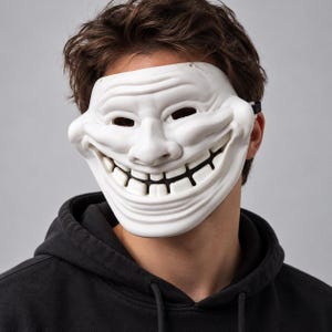 Peut inclure: Un masque en plastique blanc représentant le design du mème troll face avec des traits exagérés, incluant un large sourire édenté, des yeux plissés et des rides profondes sur le front, maintenu par une sangle élastique noire.