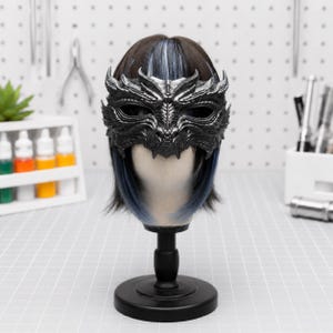 Op de afbeelding: Een donkergrijs, metallic drakenmasker met gedetailleerde schubstructuren en scherpe accenten rond de oogopeningen. Het masker wordt getoond op een etalagepophoofd met donker haar met subtiele blauwe highlights, geplaatst op een zwarte standaard.