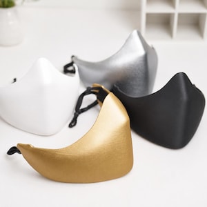 May include: A collection of four contoured face masks in solid white, metallic silver, metallic gold, and matte black. Each mask features a structured, curved design with a pointed centre seam and adjustable black elastic ear loops with toggles.