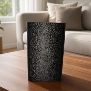 Könnte beinhalten: Eine rechteckige Vase mit einer texturierten schwarzen Oberfläche und einem dezent schimmernden Metallic-Finish. Das Objekt hat eine klare, vertikale Struktur mit scharfen Kanten und ein durchgehend dunkles Erscheinungsbild auf der Außenseite.