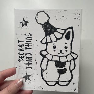 May include: A rectangular white card featuring a black ink block print of a cat wearing a clown hat and a ruffled collar. The text "SECRET THIRD THING" is printed vertically on the left side, flanked by two black stars, all enclosed in a clear sleeve.