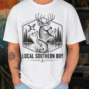 May include: A white short-sleeved t-shirt featuring a black graphic print of a deer, a jumping fish, a flying duck, mountains, and a cabin. The text LOCAL SOUTHERN BOY is printed below the illustration, accompanied by a small fishing hook icon.