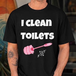 以下が含まれることがあります： 胸元に白い文字で「I CLEAN TOILETS」とプリントされた黒い半袖Tシャツ。テキストの下には、ふわふわしたヘッドのピンク色のトイレブラシのグラフィックと、右側に小さな白い輝きのアイコンがいくつか描かれています。
