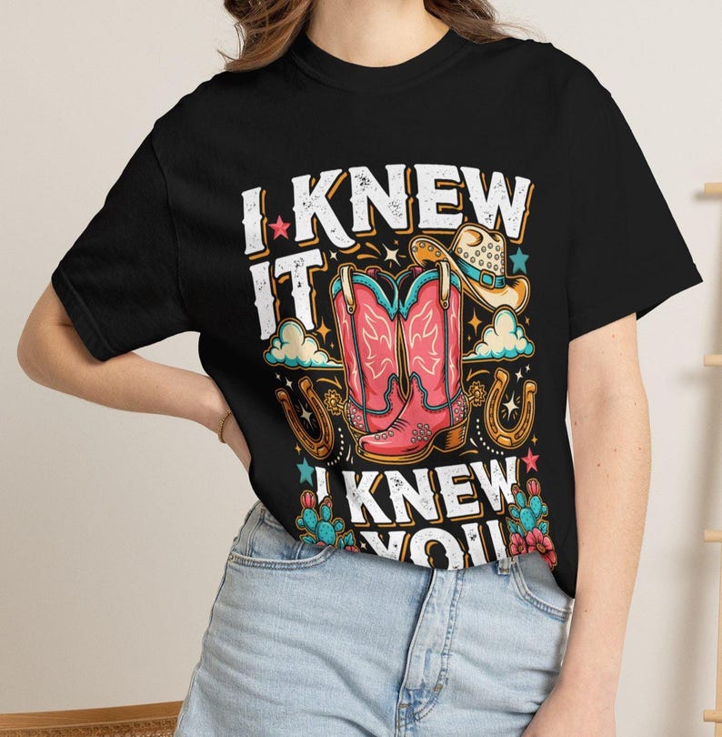 Pode incluir: Uma t-shirt preta com uma estampa gr&aacute;fica de botas de cowboy cor-de-rosa, um chap&eacute;u de cowboy bege, ferraduras e cactos. O texto "I KNEW IT" e "I KNEW YOU" est&aacute; impresso em letras brancas desgastadas em torno da ilustra&ccedil;&atilde;o central.