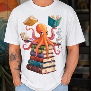 Puede incluir: Una camiseta blanca de manga corta con un gráfico de un pulpo naranja posado sobre una pila de cinco libros coloridos. El pulpo sostiene dos libros abiertos con sus tentáculos y una taza de café humeante sobre un platillo a la derecha.