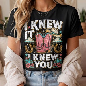 Pode incluir: Uma t-shirt preta com uma estampa gr&aacute;fica contendo o texto I KNEW IT I KNEW YOU. O design inclui um par de botas de cowboy cor-de-rosa, um chap&eacute;u de cowboy bege, duas ferraduras douradas, nuvens azuis, pequenas estrelas e cactos verdes.