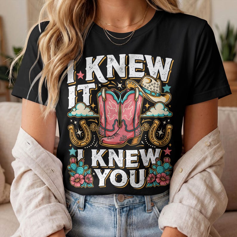 Pode incluir: Uma t-shirt preta com uma estampa gr&aacute;fica contendo o texto I KNEW IT I KNEW YOU. O design inclui um par de botas de cowboy cor-de-rosa, um chap&eacute;u de cowboy bege, duas ferraduras douradas, nuvens azuis, pequenas estrelas e cactos verdes.