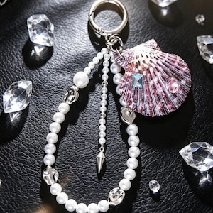 May include: A keychain featuring a silver-toned ring, a loop of white faux pearls, and a purple seashell charm. The shell is decorated with a small rectangular blue gem and a round pink gem. A silver spike charm hangs from the center of the pearl loop.
