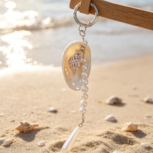 May include: A keyring featuring a silver-toned metal ring attached to a natural seashell. Inside the shell is a smaller spiral shell, a strand of white spherical beads, and a single white teardrop-shaped pendant hanging from the metal chain.