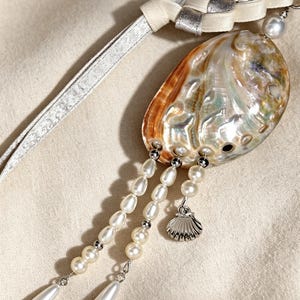 May include: A bag charm and keychain featuring an iridescent abalone shell, white freshwater pearls, and a small silver-toned metal seashell charm. The piece includes a braided cream and silver ribbon strap with the text Etsy Seller Handmade Shell Bag Charm & Keychain.