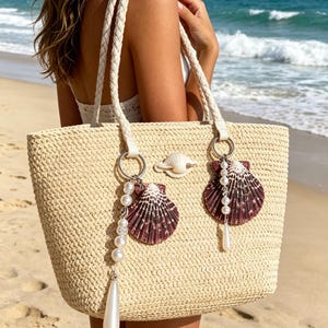 May include: A beige woven straw tote bag featuring two braided handles and silver-tone metal rings. The bag is decorated with two dark purple scallop shells, several white faux pearl beads, and a single white teardrop-shaped pendant.