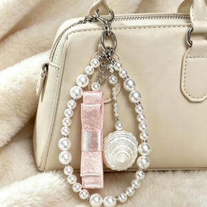 May include: A cream-colored handbag featuring a decorative charm attached to the handle. The charm consists of a teardrop-shaped loop of white faux pearls, a light pink satin ribbon bow, and a single white seashell pendant on a silver chain.