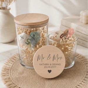 May include: Two clear glass jars with wooden lids, filled with dried flowers and Euro banknotes. A wooden coaster with "Mr & Mrs", names, date, and a heart is in front. The jars sit on a burlap mat.