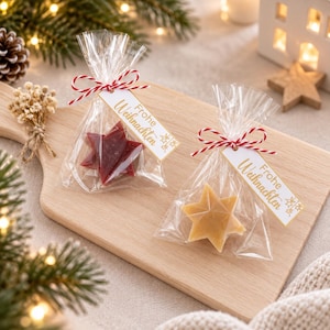 May include: Two clear cellophane bags each containing a single star-shaped item, one dark red and one light tan. Each bag is tied with red and white baker's twine and features a white rectangular tag with the gold text Frohe Weihnachten.