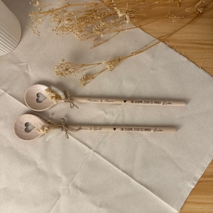 May include: Two wooden spoons with heart-shaped cutouts and dried floral accents. Each spoon is engraved with a name and the phrase "DIE GEHEIME ZUTAT IST IMMER Liebe". Tied with twine, they rest on a cream-colored surface.