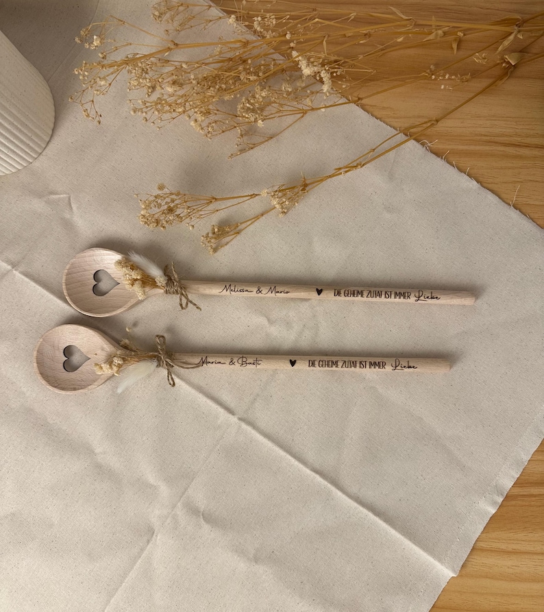 May include: Two wooden spoons with heart-shaped cutouts and dried floral accents. Each spoon is engraved with a name and the phrase "DIE GEHEIME ZUTAT IST IMMER Liebe". Tied with twine, they rest on a cream-colored surface.