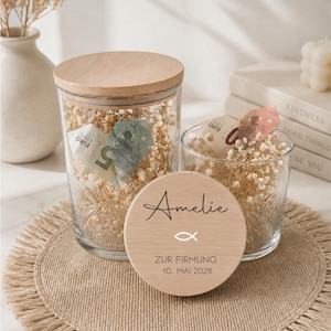 May include: Two clear glass jars with wooden lids, filled with dried flowers and currency. A wooden disc with the name "Amelie", a fish symbol, and text "ZUR FIRMUNG 10. MAI 2026" is in front of the jars. The jars sit on a burlap mat.