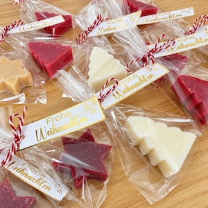 May include: Multiple wax melts shaped like Christmas trees and stars in red, white, and tan colors. Each is individually wrapped in clear plastic, tied with red and white baker's twine, and features a white tag with the text Frohe Weihnachten.