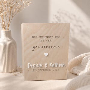 Könnte beinhalten: Eine rechteckige Box aus hellem Holz mit dem deutschen Text "DER SCHÖNSTE WEG IST DER gemeinsame" und einem kleinen weißen Herzsymbol darunter. Die Namen "Dennis & Kathrin" sowie das Datum "22. SEPTEMBER 2017" sind in Weiß abgebildet.
