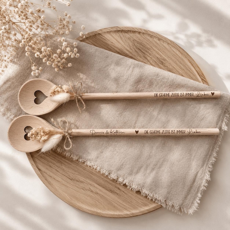 May include: Two wooden spoons with heart-shaped cutouts and dried floral accents. The spoons have text that reads "Dennis & Kathrin" and "DIE GEHEIME ZUTAT IST IMMER Liebe" with a heart symbol. They are tied with twine.