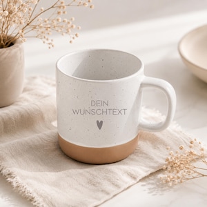 May include: A ceramic mug with a speckled white upper section and a solid tan base. The front features the text DEIN WUNSCHTEXT in grey, positioned above a small grey heart icon. The mug has a curved handle and a smooth, cylindrical shape.