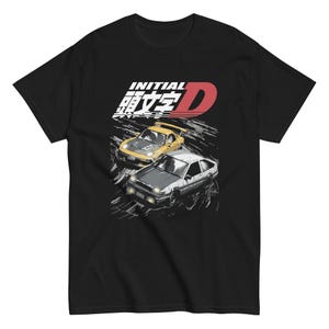 Könnte beinhalten: Ein schwarzes T-Shirt mit kurzen Ärmeln und einem Grafikdruck, der einen gelben und einen weißen Sportwagen zeigt, die auf einer Straße driften. Über den Autos steht der Text INITIAL sowie japanische Schriftzeichen und ein rotes D.