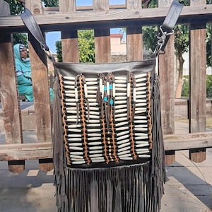 May include: A dark brown leather shoulder bag with a fringed bottom and decorative bone and bead accents. The bag has a rectangular shape with a long strap and is hanging on a wooden fence.