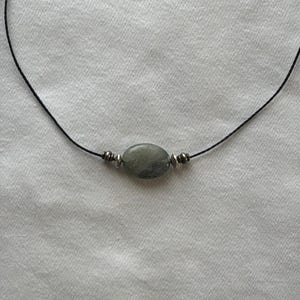 May include: A necklace featuring a single oval-shaped gray stone bead centered on a thin black cord. The stone is flanked on each side by two small, metallic silver-toned spacer beads, creating a simple and balanced design for this piece of jewelry.