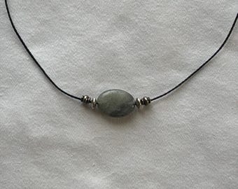 Puede incluir: Un collar que presenta una cuenta de piedra gris de forma ovalada centrada en un cordón negro fino. La piedra está flanqueada a cada lado por dos pequeñas cuentas espaciadoras de tono plateado metálico, creando un diseño sencillo y equilibrado.