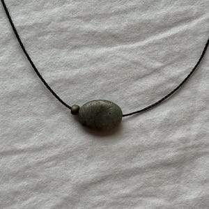 Puede incluir: Un collar que consiste en un cordón negro fino con un colgante de piedra gris de forma ovalada y una pequeña cuenta redonda de tono metálico situada justo a la izquierda de la piedra, formando una pieza de joyería sencilla y discreta.