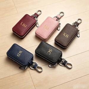May include: Five rectangular leather key cases with zippered closures and silver-toned metal swivel clips. The cases are available in burgundy, light pink, brown, navy blue, and black, each featuring gold-embossed initials: LBJ, MJ, KB, WD, and SC.