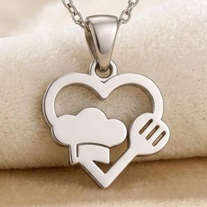 May include: A silver-colored pendant necklace featuring a heart-shaped design with a chef's hat, cloud, and spatula. The necklace hangs on a delicate chain, perfect for culinary enthusiasts.