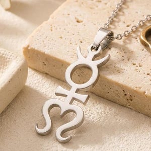 May include: A silver pendant necklace displaying the astrological symbol for Mercury. The pendant has a circular element, a crescent shape, and a heart-like shape. The necklace is presented on a textured, light-coloured surface. The chain is a delicate design.