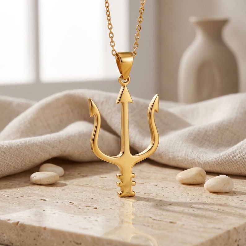 A polished silver or rose gold Neptune Trident Necklace, featuring a three-pronged spear pendant, rests on a light-colored, textured stone surface. The trident design is sleek and minimalist, reflecting mythological power and ocean themes. A delicate matching cable chain connects to the pendant, with a subtle background of dried wheat stalks and soft, neutral tones. This sterling silver piece is a symbolic gift for strength and protection, capturing the essence of the sea god's iconic weapon.
