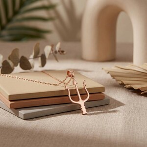 Puede incluir: Un collar con colgante de tridente en oro rosa. El colgante es un dise&ntilde;o de tenedor de tres puntas, sujeto a una delicada cadena. El collar descansa sobre una pila de bloques cuadrados de color neutro, con un fondo suave y borroso.
