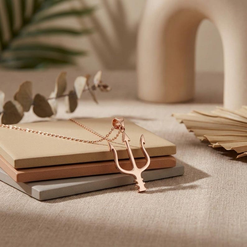 Puede incluir: Un collar con colgante de tridente en oro rosa. El colgante es un dise&ntilde;o de tenedor de tres puntas, sujeto a una delicada cadena. El collar descansa sobre una pila de bloques cuadrados de color neutro, con un fondo suave y borroso.