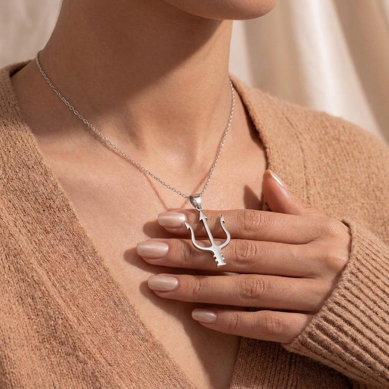 Puede incluir: Un collar plateado con un colgante de tridente. El colgante es un dise&ntilde;o de tenedor de tres puntas. El collar lo lleva una persona con un su&eacute;ter color beige.