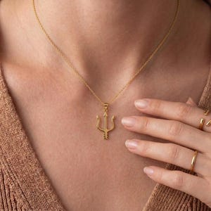 Puede incluir: Un collar dorado con un colgante de tridente. El colgante es un dise&ntilde;o de tenedor de tres puntas, suspendido de una delicada cadena. El collar se lleva alrededor del cuello, y la imagen muestra una vista de cerca de la joya.